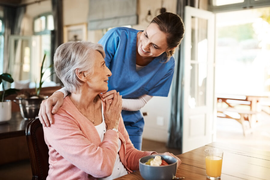 Healthcare, elderly woman with nurse with breakfast at her home and at the table in living room. Support or communication, caregiver and conversation with medical person with senior or old female