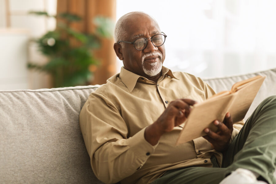 Senior African American Man Reading Book Sitting On Couch At Home, Wearing Eyeglasses. Retired Male Enjoying Reading New Novel Or Business Literature On Weekend. Retirement Leisure Concept